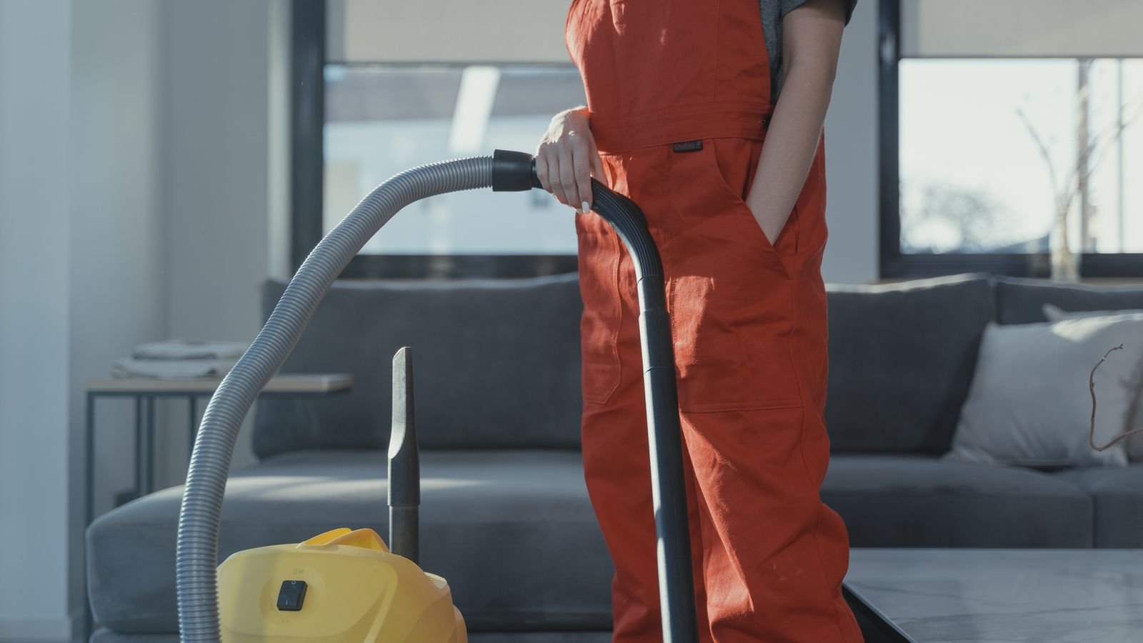 Cleaner in overalls vacuuming a floor beside a sofa