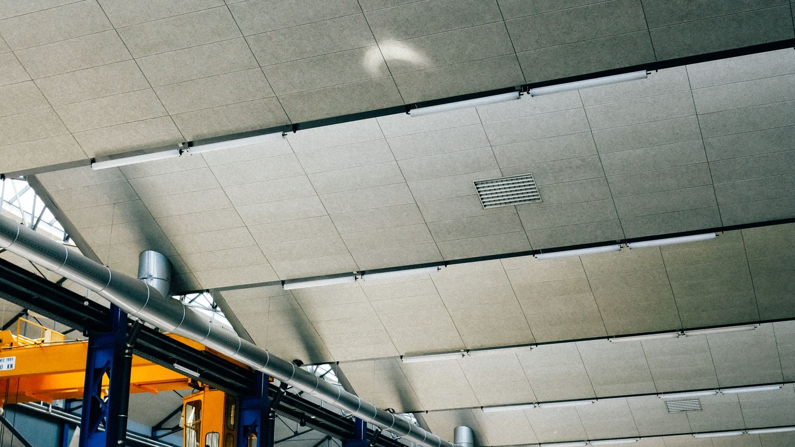 The panelled ceiling of a large industrial hall with strip lights, pipework and blue steel columns.