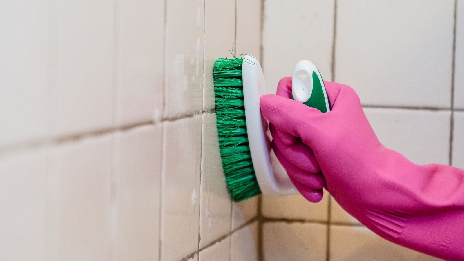 Pink gloved hand scrubbing discoloured grout on white bathroom tiles above a bath