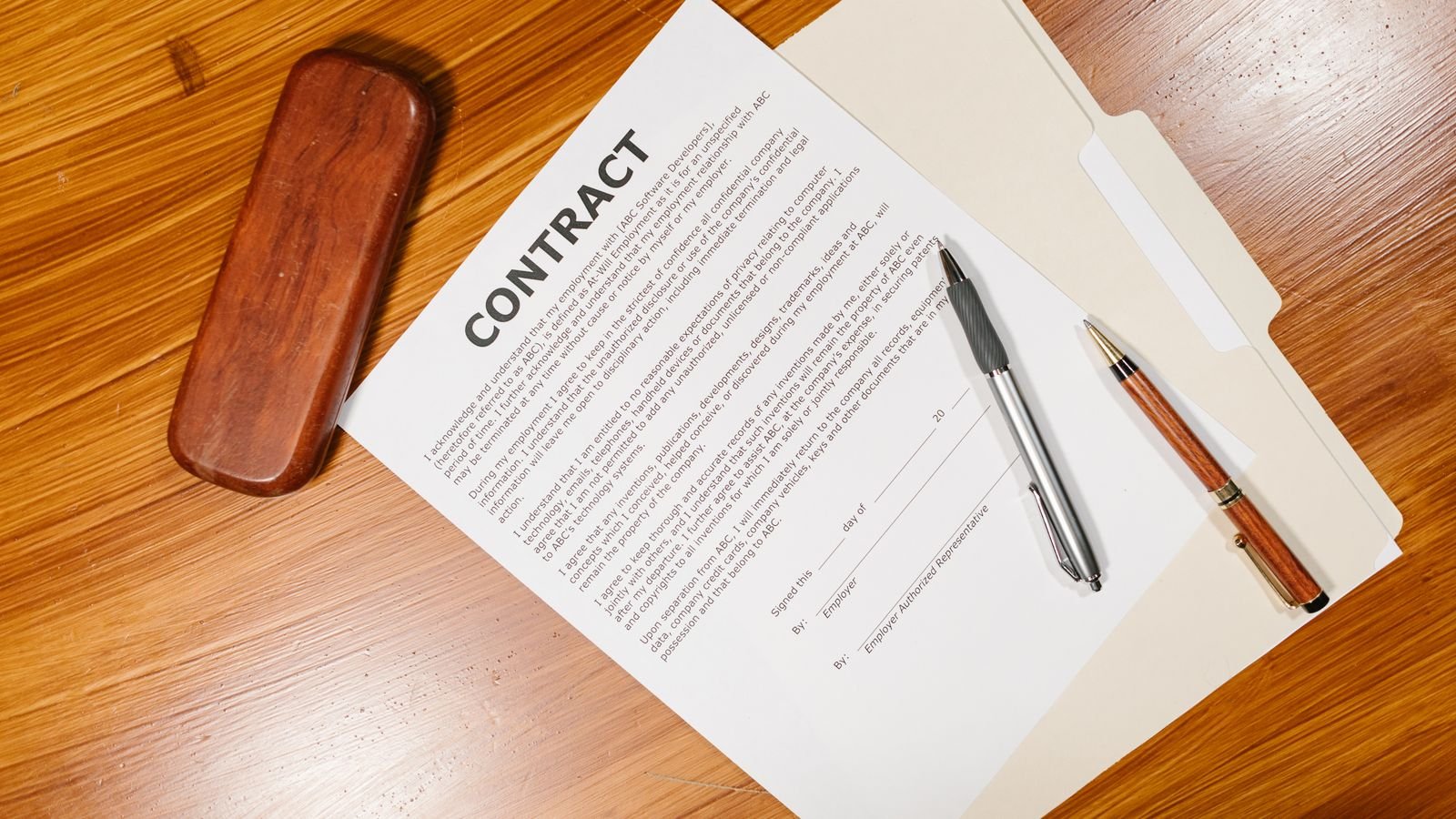A contract and two pens lie on a folder on a wooden desk, beside a wooden pen case.