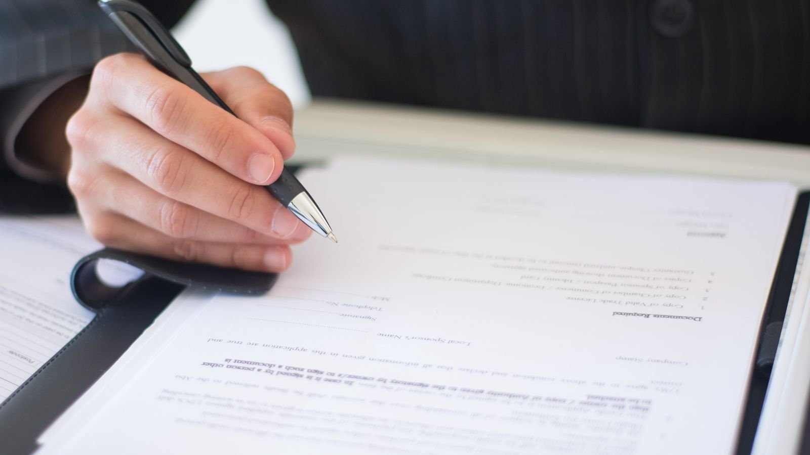 A hand signs a form held in a black folder on a desk. A hand signs a form held in a black folder on a desk.