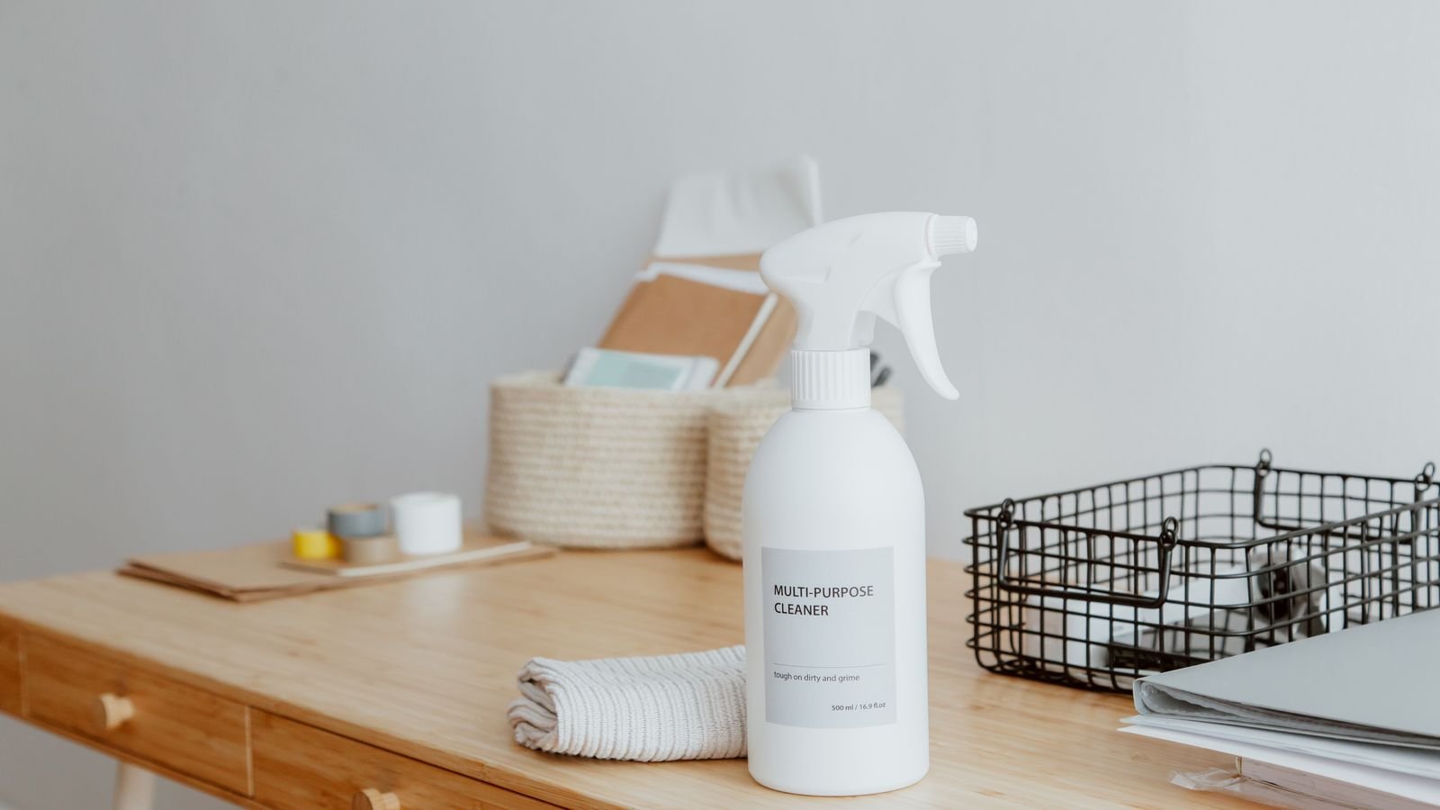 A white spray bottle of multi-purpose cleaner on a wooden desk beside a folded cloth and a wire tray of papers.