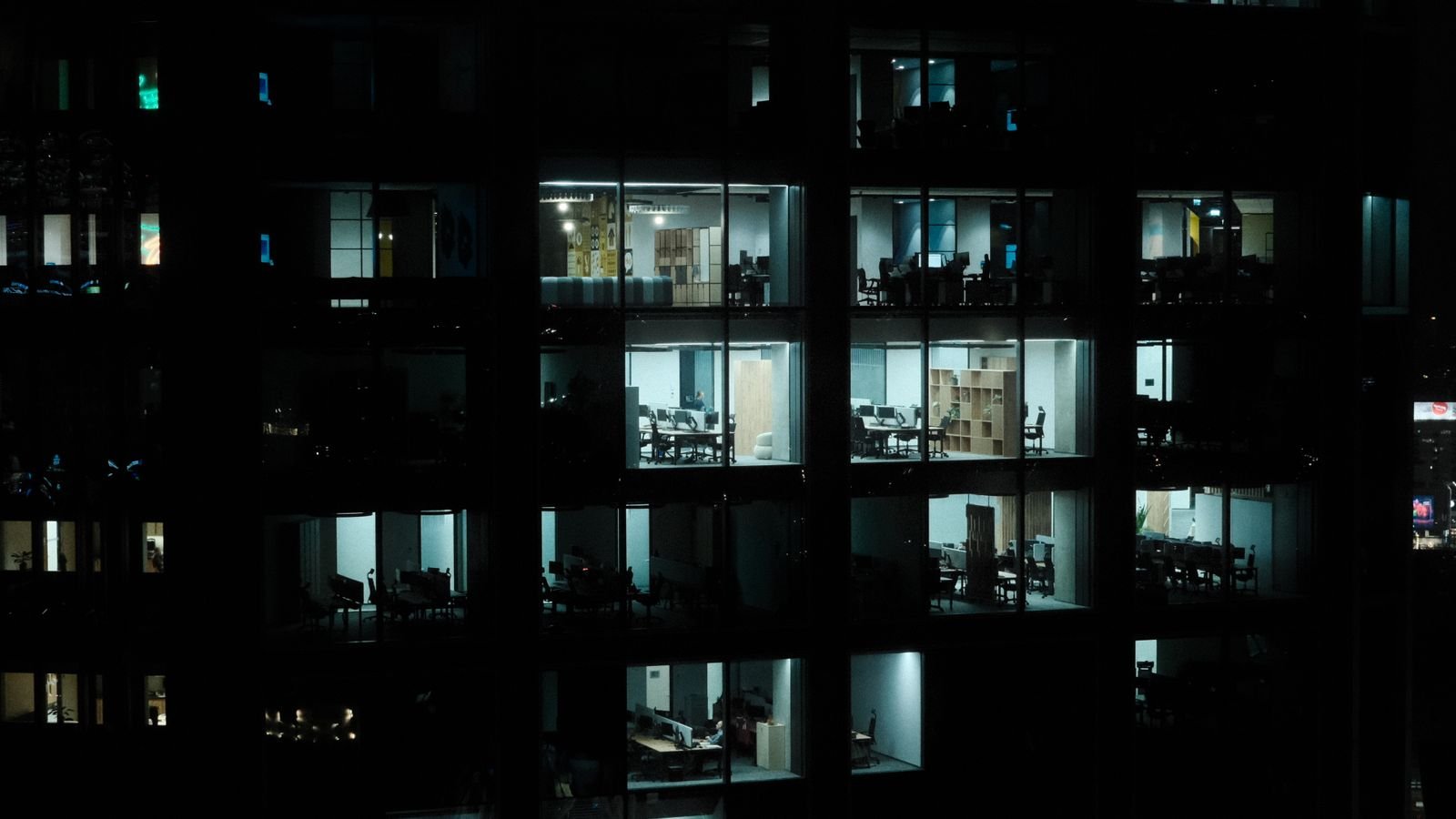 Office tower at night with a few lit floors showing empty desks and meeting rooms