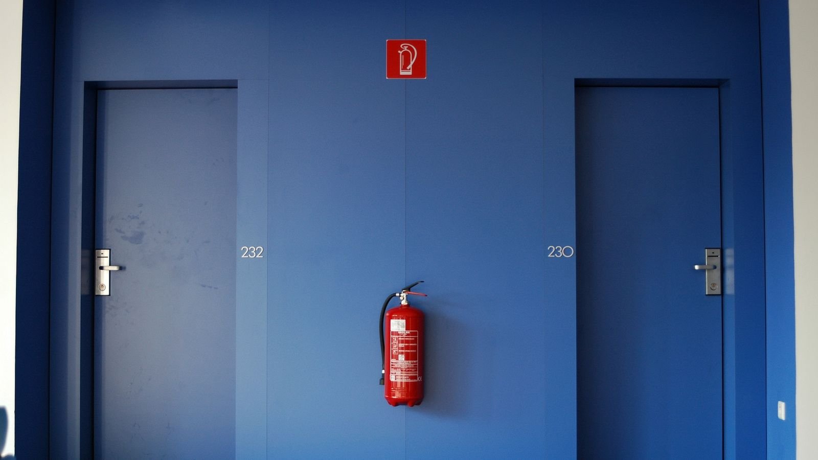 A red fire extinguisher hangs on a blue wall between two doors, with a fire extinguisher sign above it. A red fire extinguisher hangs on a blue wall between two doors, with a fire extinguisher sign above it.