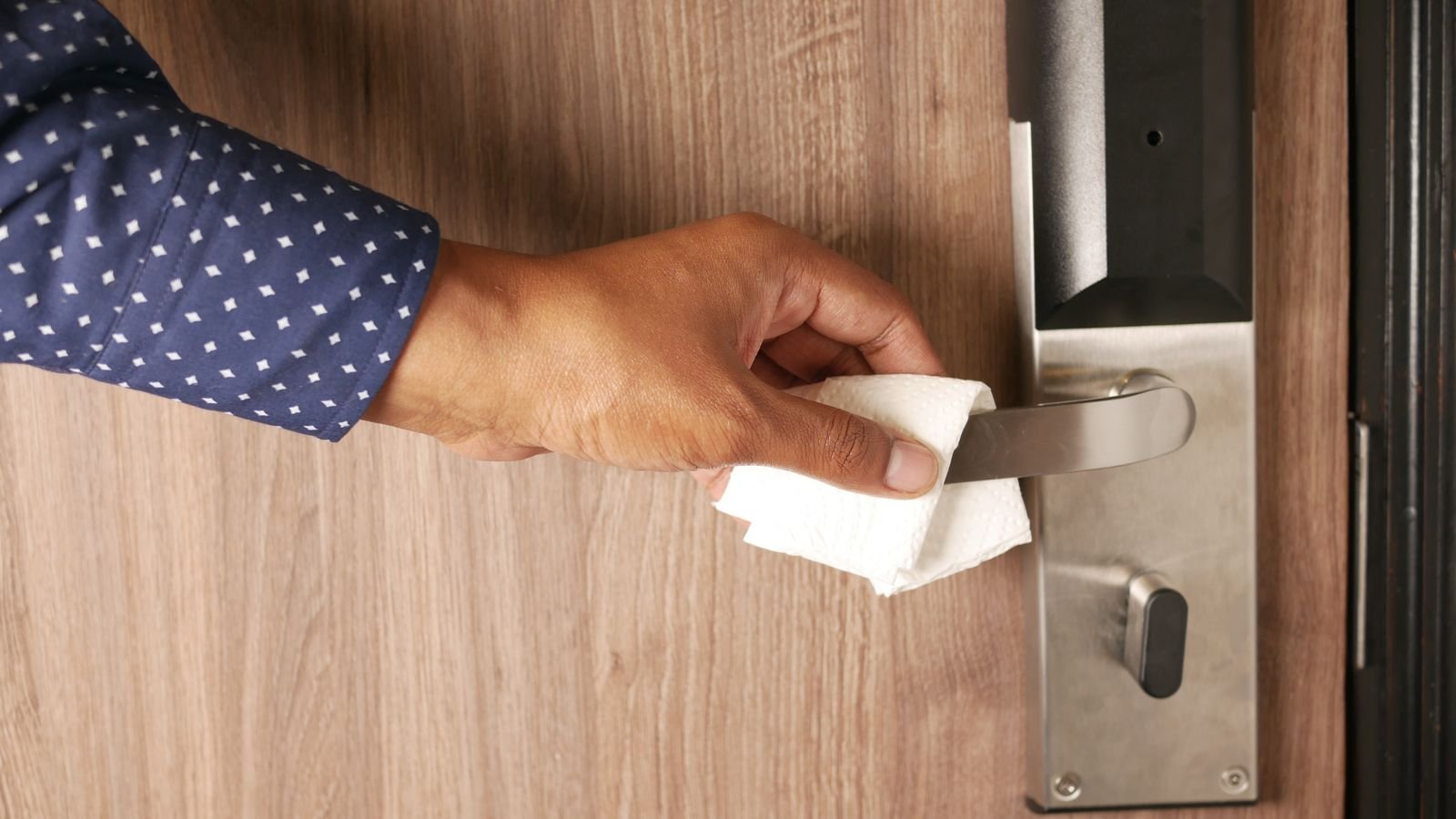 A hand in a blue shirt wipes a stainless steel lever door handle with a disinfectant wipe