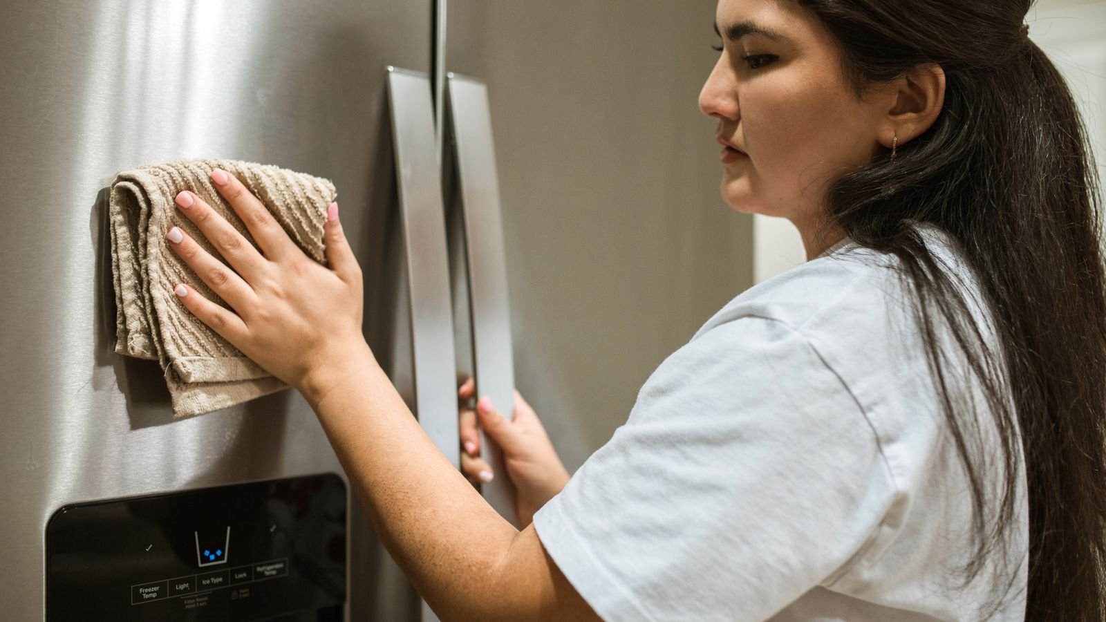 A woman in a white t-shirt wipes the handle of a stainless steel fridge door with a cloth. A woman in a white t-shirt wipes the handle of a stainless steel fridge door with a cloth.