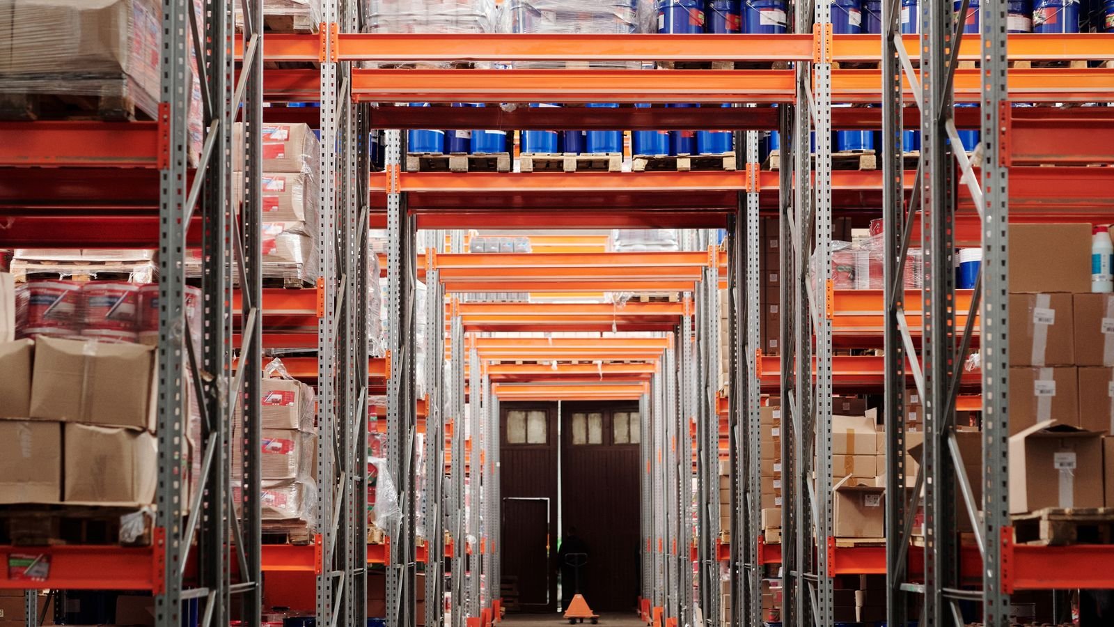 Tall orange warehouse racking with pallets stacked high on both sides of a long aisle. Tall orange warehouse racking with pallets stacked high on both sides of a long aisle.