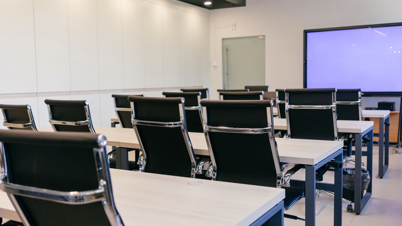 Black office chairs along white desks in a bright training room with a wall screen.