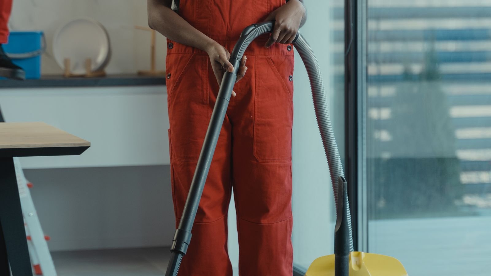 A cleaner in orange overalls holds the hose of a wet and dry vacuum beside a glass wall in a bright office.